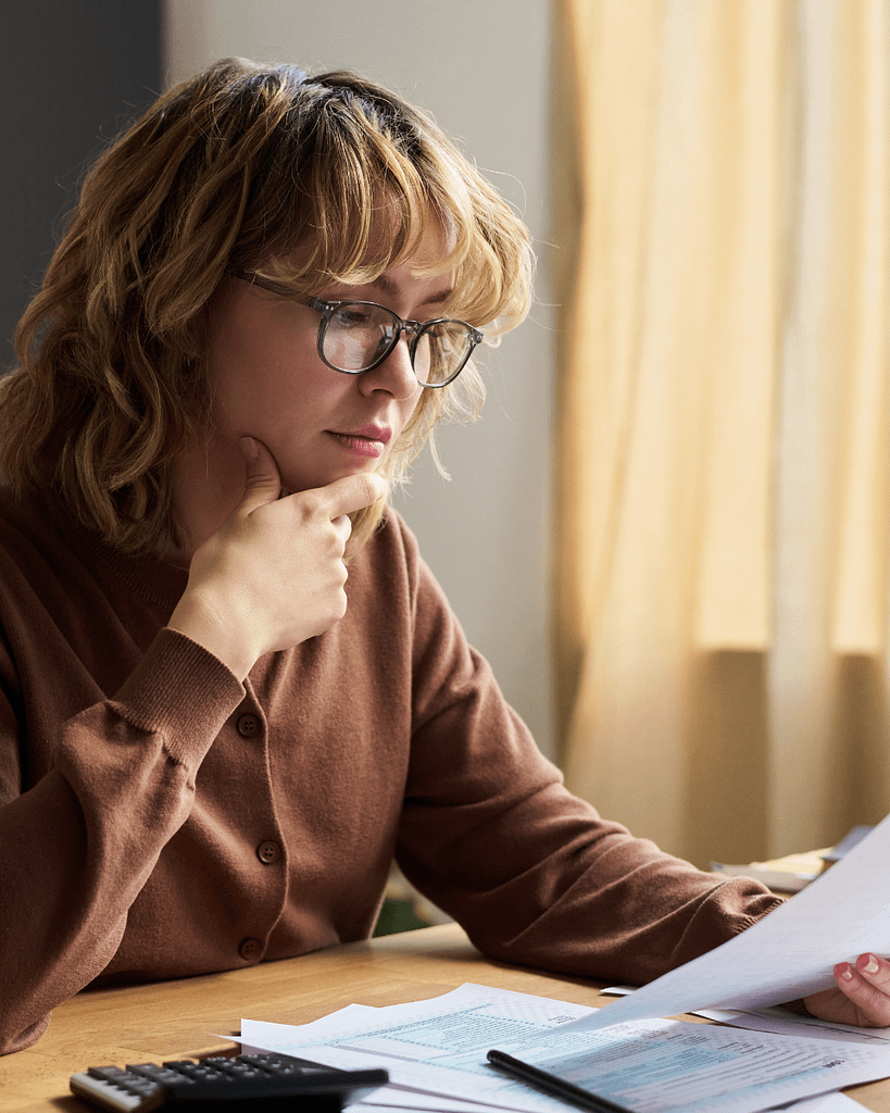 Woman sitting at a table reviewing bills and financial paperwork while looking thoughtful and financially overwhelmed.
